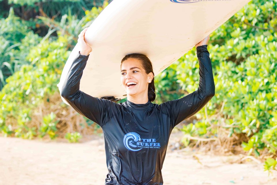 Lachende Surferin am Strand trägt ihr Board auf dem Kopf